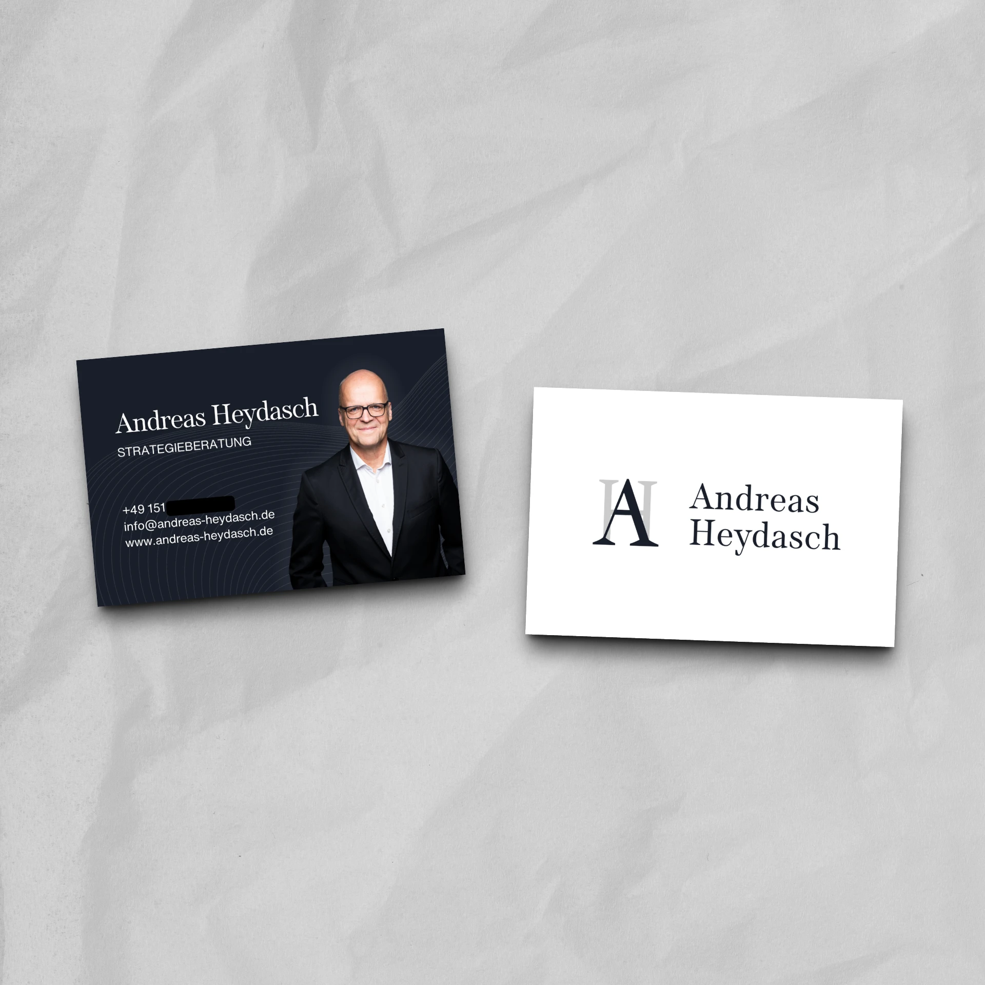 Zwei Visitenkarten für Andreas Heydasch zeigen elegantes Print-Design. Die linke Karte ist dunkel und enthält sein Foto, seinen Namen, seinen Titel, seine Kontaktdaten und seine Website. Die rechte Karte zeigt AH und seinen Namen in schwarzer Serifenschrift. Die Visitenkarten sind auf eine graue Oberfläche gedruckt.